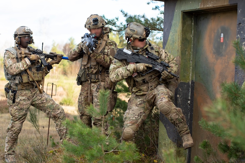 A soldier in camouflage kicks down a door as two others stand at the ready.