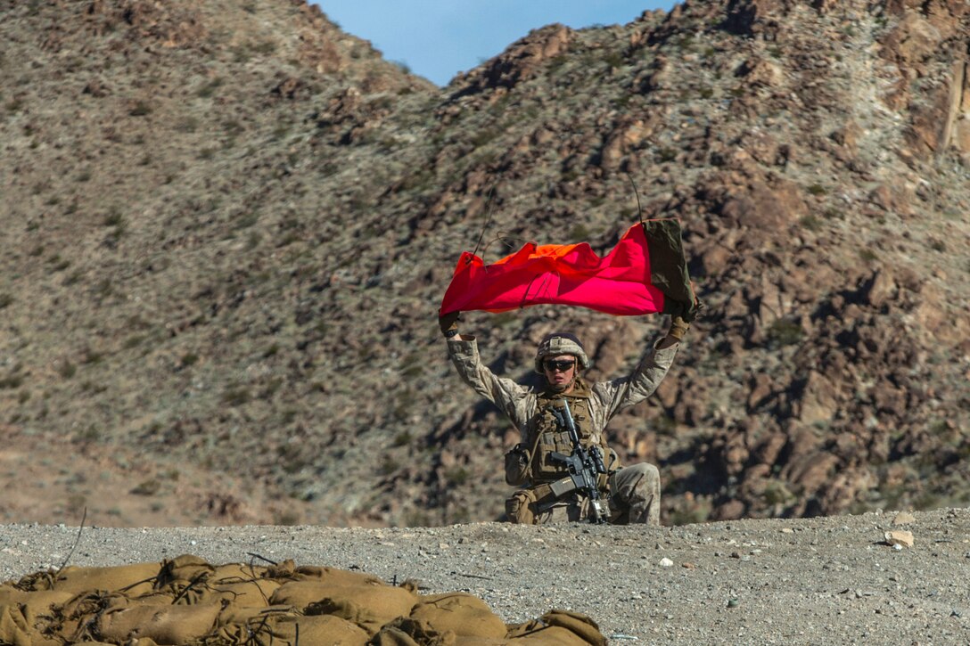 A Marine holds a red flag over his head while standing in front of mountains in desert terrain.