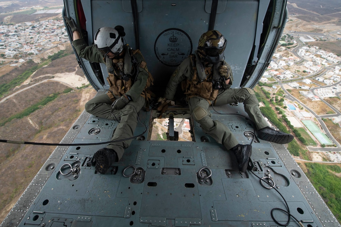 Two men sit in a helicopter as it flies above land.