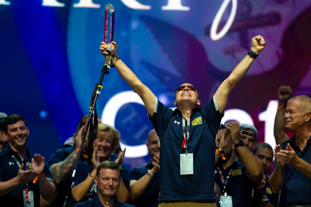 A sailor raises up his arms holding an award with a group of people clapping in the background.