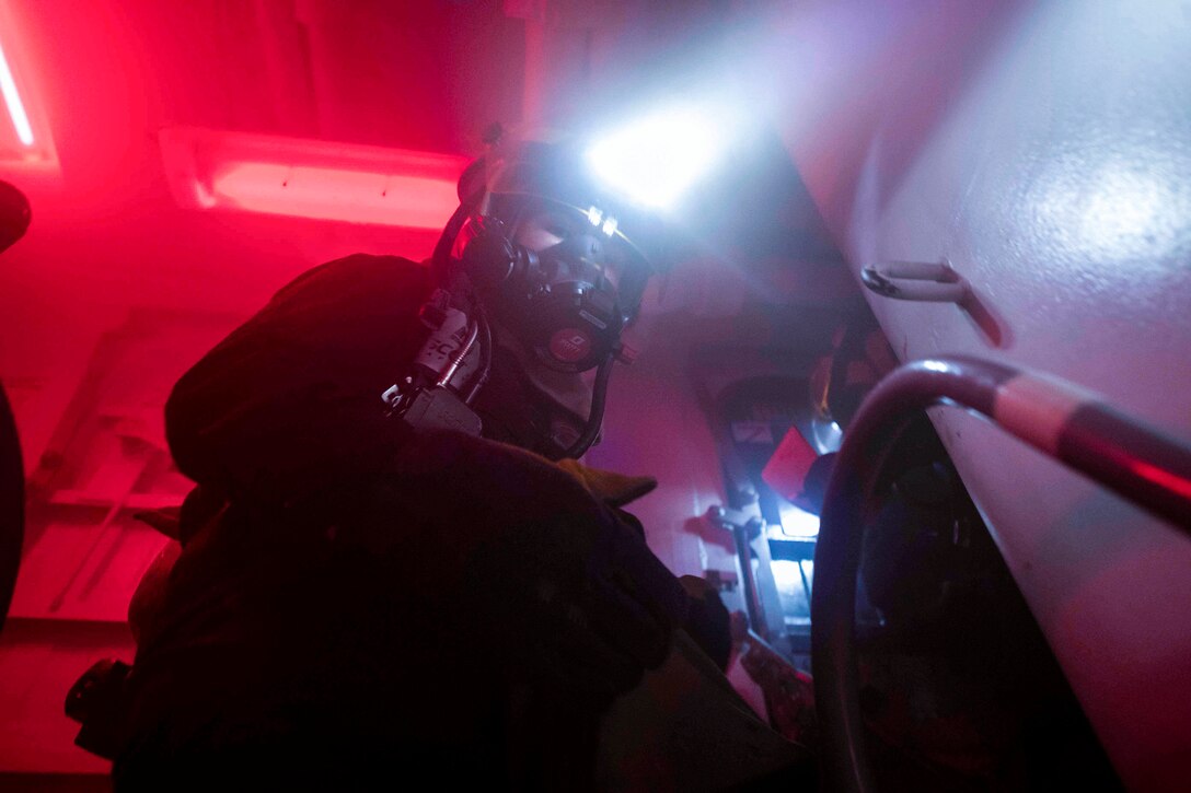 A sailor wearing a helmet with a light at the top walks through a small space.