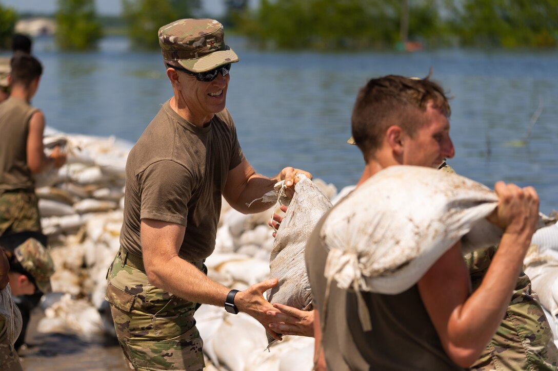 A general and several guardsmen place sandbags near a river’s edge.