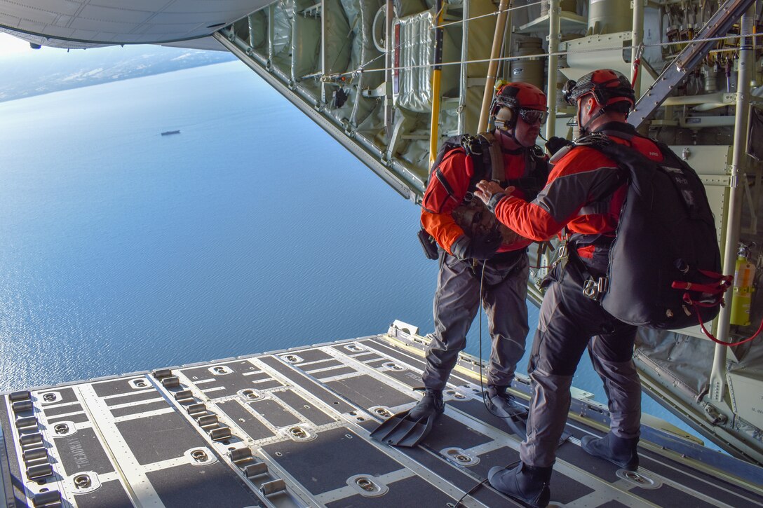 Two airmen stand on the open end of a military jet in flight as they prepare to jump.