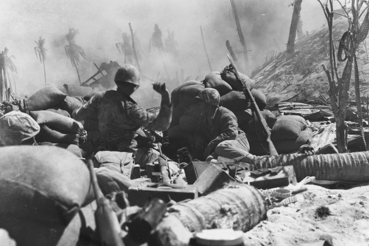 Marines hide behind sandbags during heavy combat beside a hill; one Marine’s hand is cocked backward, ready to launch a grenade into the distance. Sand is in the foreground; palm trees are in the background.