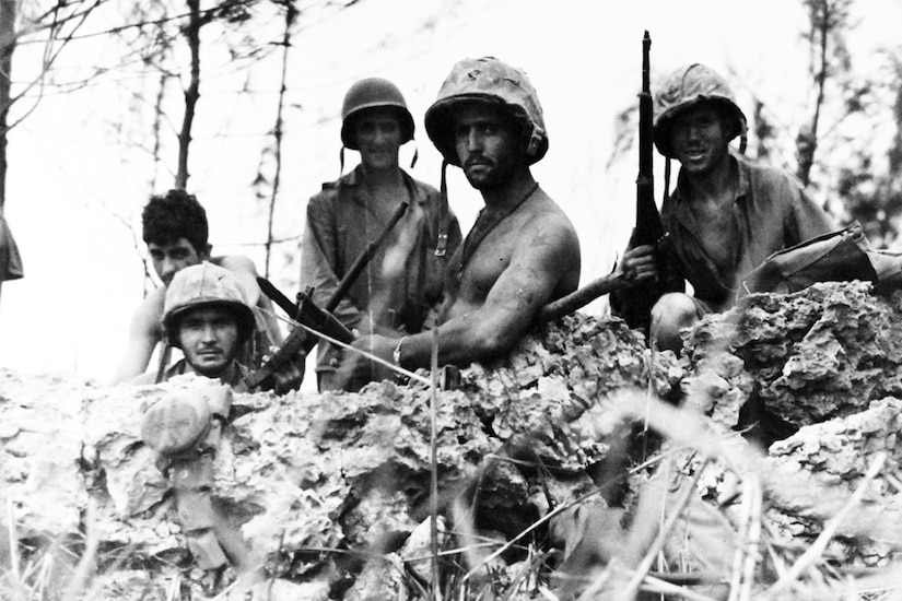 Five Marines wearing combat helmets and holding guns stand in a foxhole with sparse trees in the background.