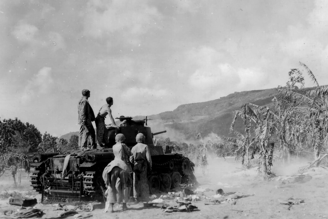 Four service members, two of whom are standing on a tank, look into the distance toward an island hill. Palm trees and sand surround them.