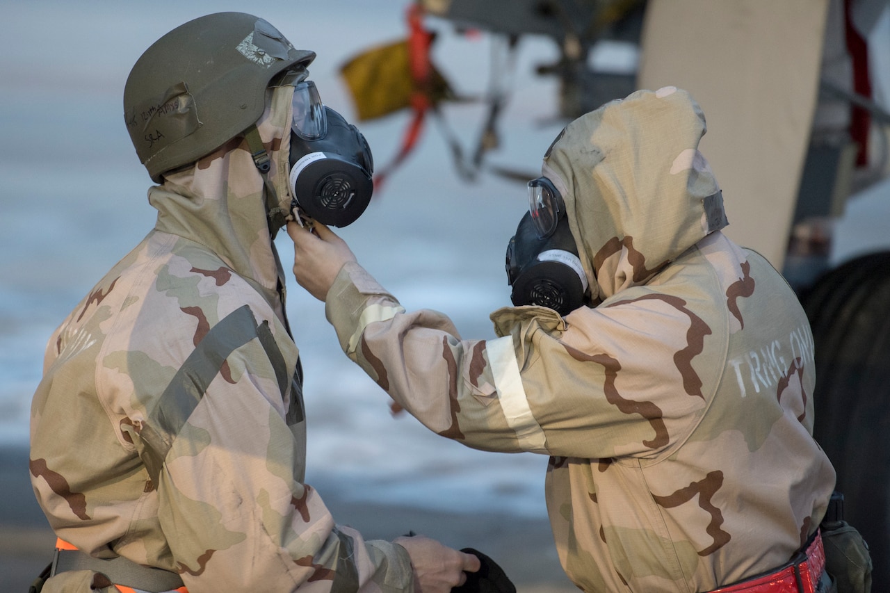 Two service members in chemical warfare gear check each other’s gas masks.
