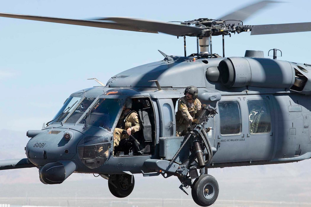 An airman holding a large gun leans out the side of a helicopter in the air.