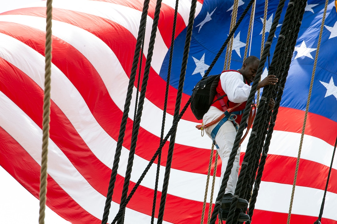 A sailor climbs down the rigging of the USS Constitution while a large American flag waves behind him.