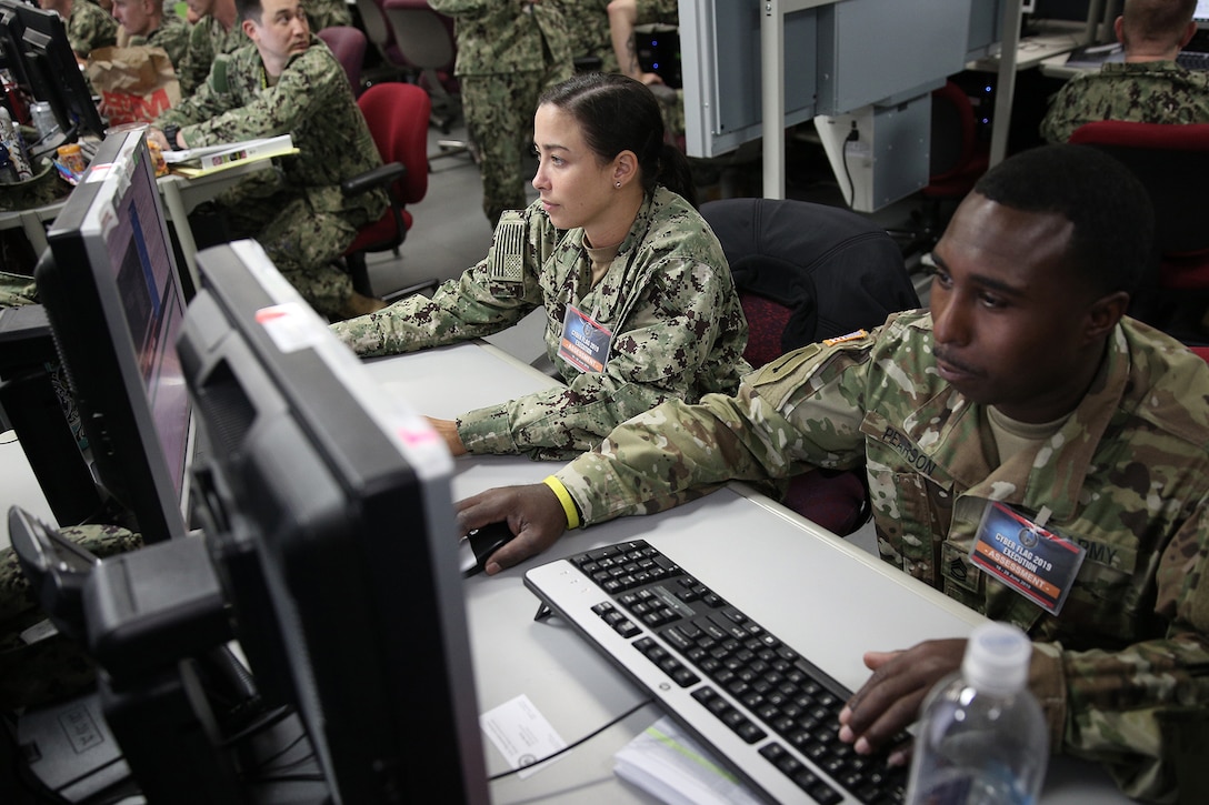 Two uniformed military personnel, one with the U.S. Navy, and one with the U.S. Army, operate computers.
