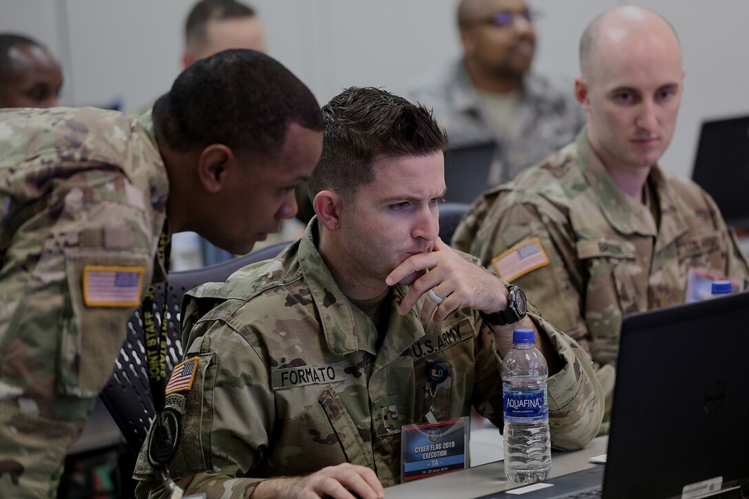 A U.S. service member sits at a desk operating a computer.  Another service members stands next to him.