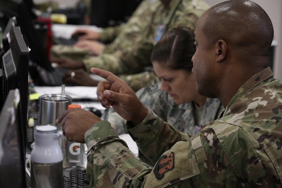 In a busy room full of computer screens, a uniformed man points at a screen.