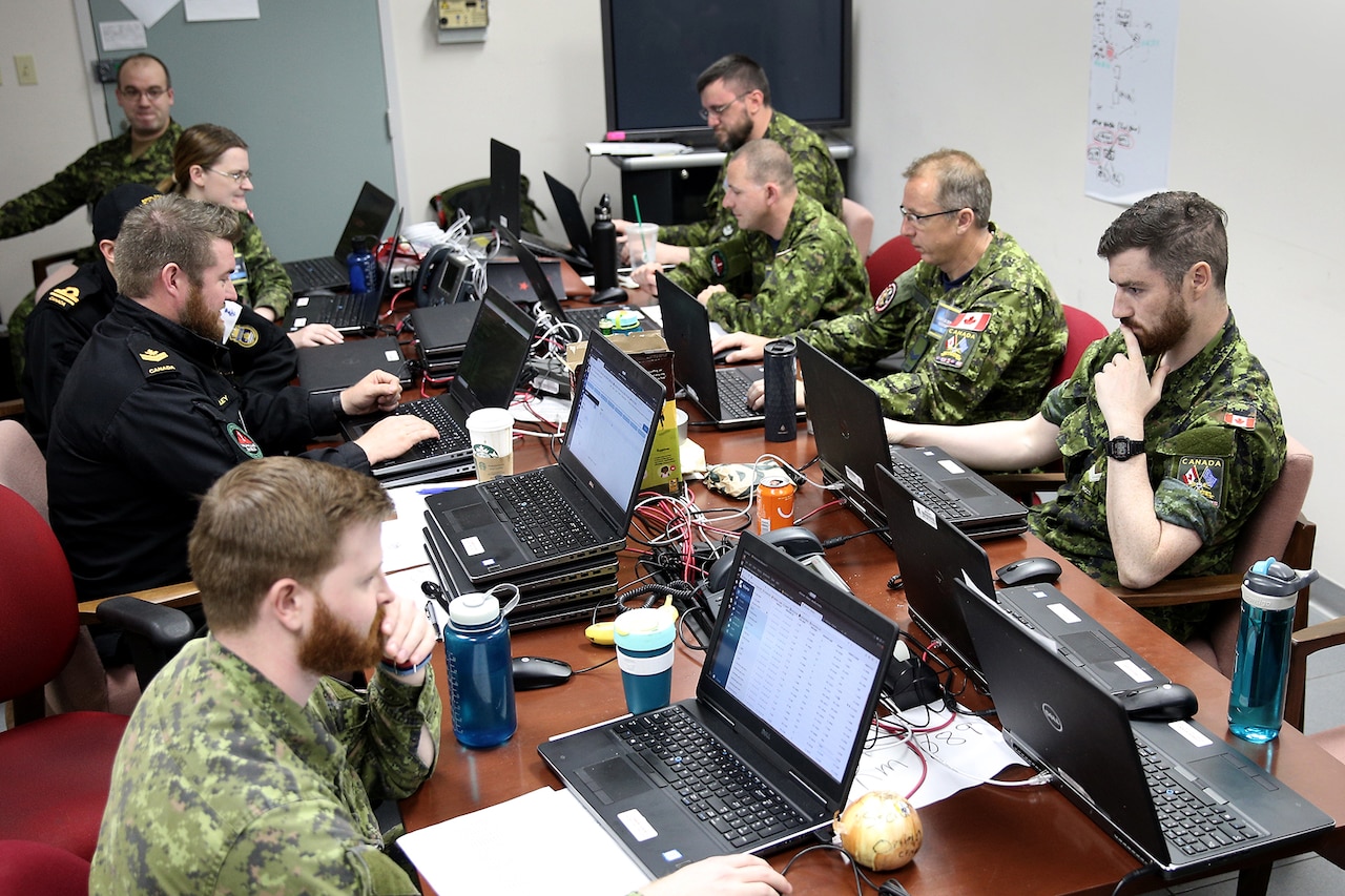 Nine uniformed military personnel, in Canadian military uniforms, sit on either side of a long table and look at laptop computer screens.