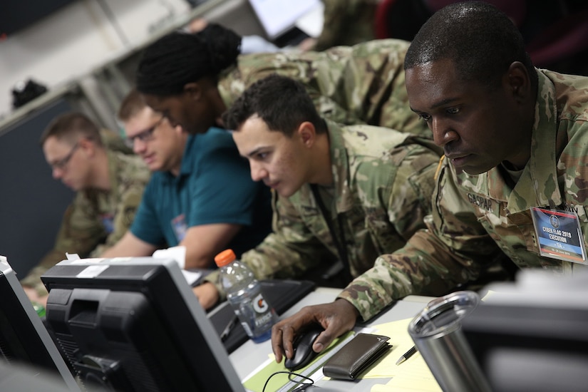 A mix of military and civilian personnel sit at a table and operate computers.  One female service member is standing and looking over the shoulder of the only civilian computer operator.