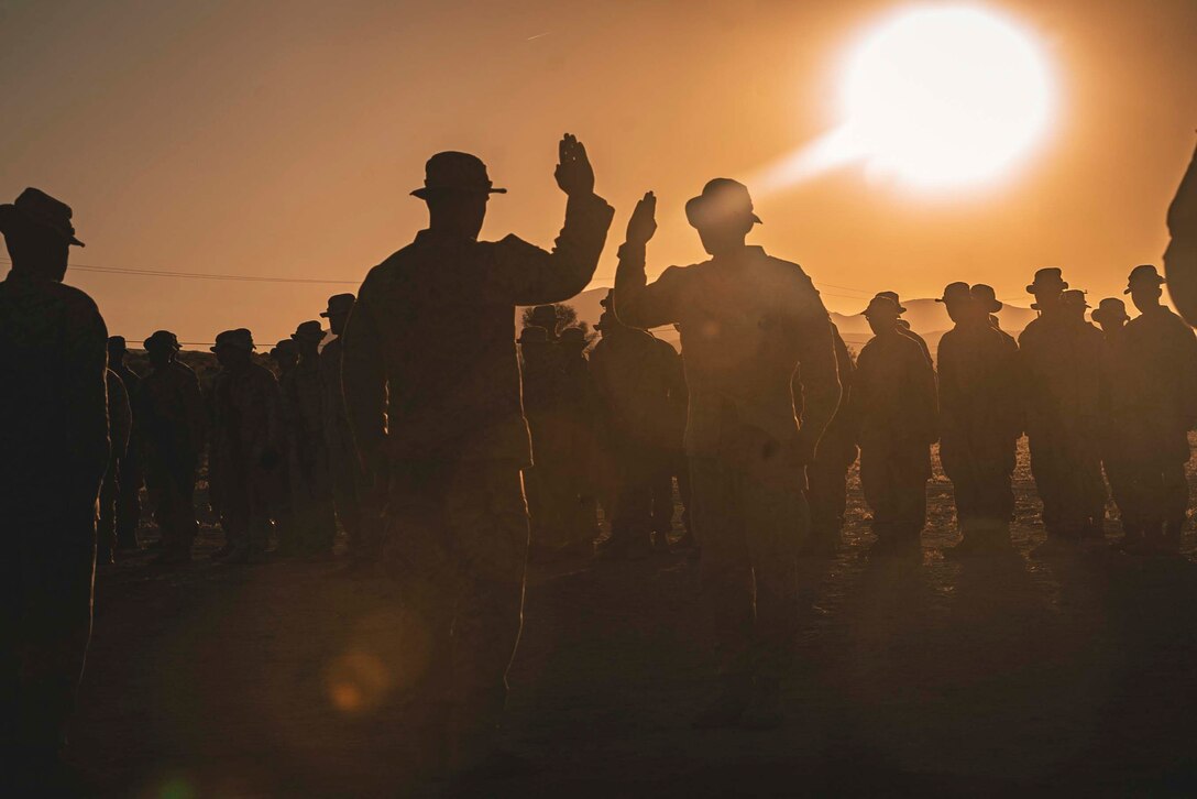 Two Marines hold up their right hands while a large group of Marines surround the two.