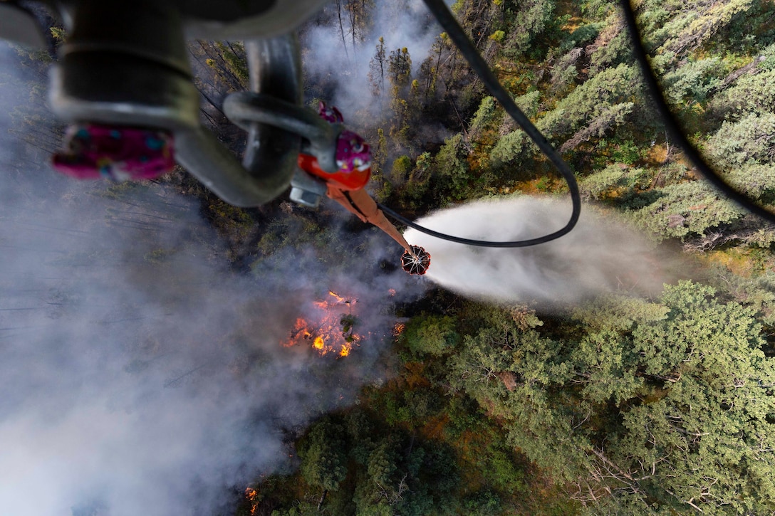 A bucket dangles from a helicopter above a forest wildfire.