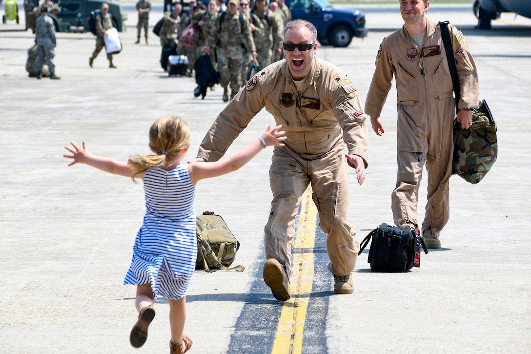 A smiling airman opens his arms as a child runs toward him with open arms on a flightline.