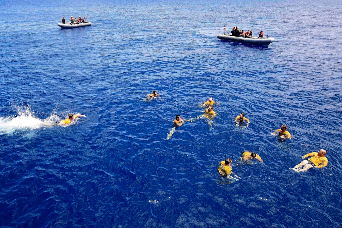 A group of sailors swim in the ocean while two inflatable boats stand watch.