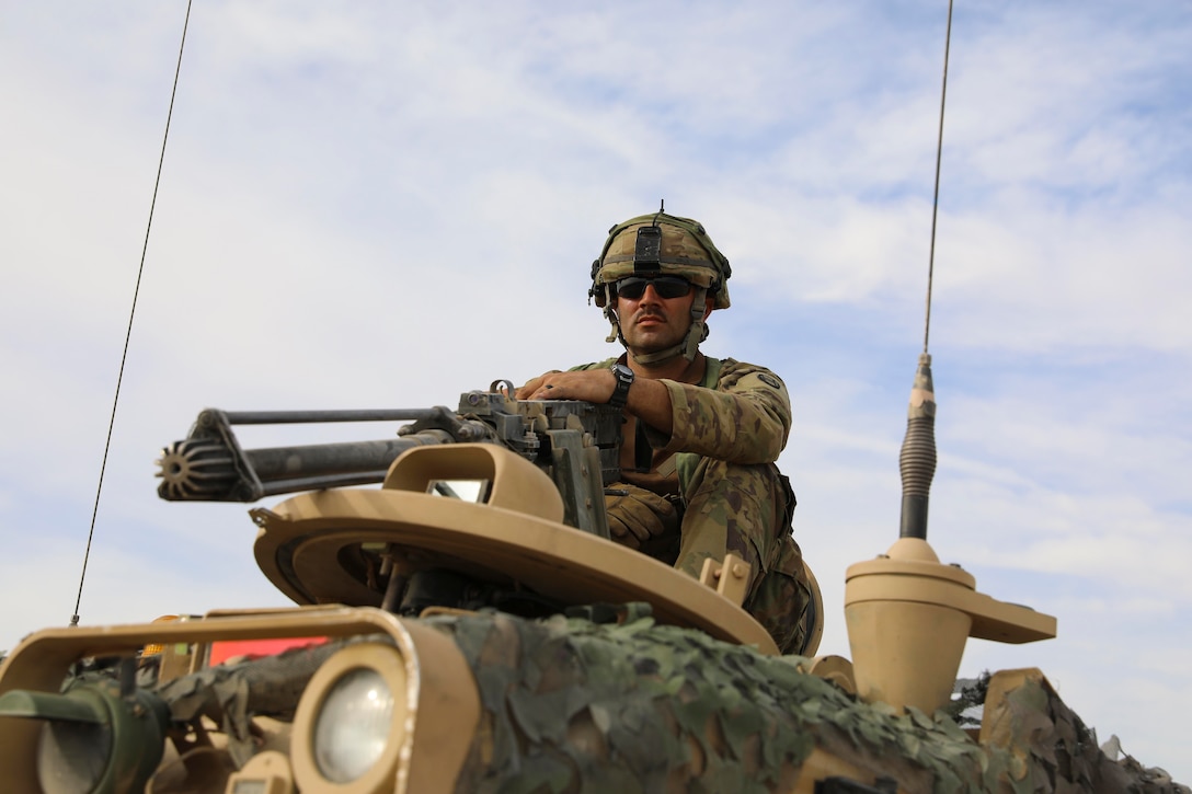 A soldier rides on top of a military vehicle while holding the machine gun attached to the top.