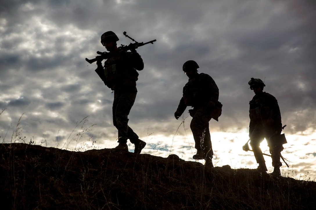 Three Marines, shown in silhouette, walk up hilly terrain, one with a weapon on his shoulders.