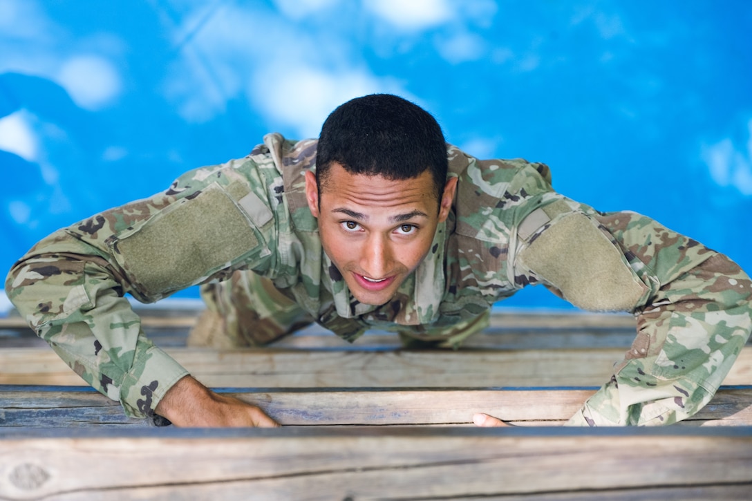 An Army ROTC cadet, shown from above, climbs a wood ladder against a vivid aqua background.