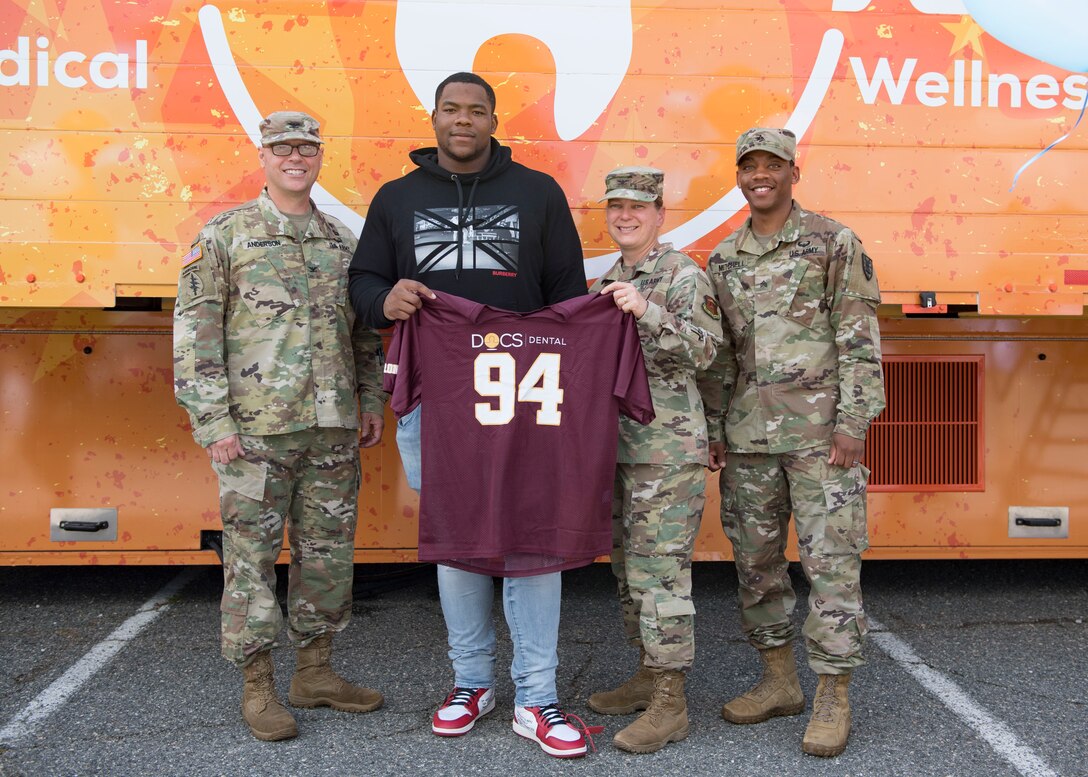 National Football League player Daron Payne, Washington Redskins defensive tackle, and Fort Eustis personnel hold up a football jersey during the grand opening of the Dentrust Optimized Care Solutions mobile treatment facility at Joint Base Langley-Eustis, Virginia, July 8, 2019.