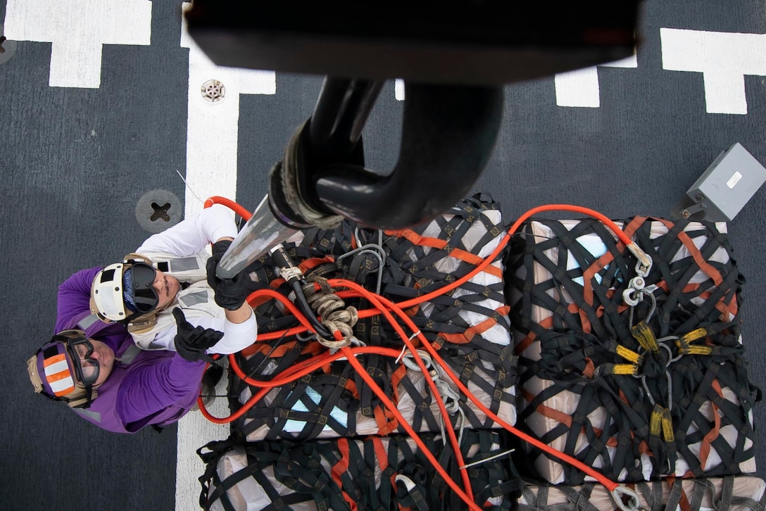 From above, two sailors try to latch a a pole onto a helicopter.