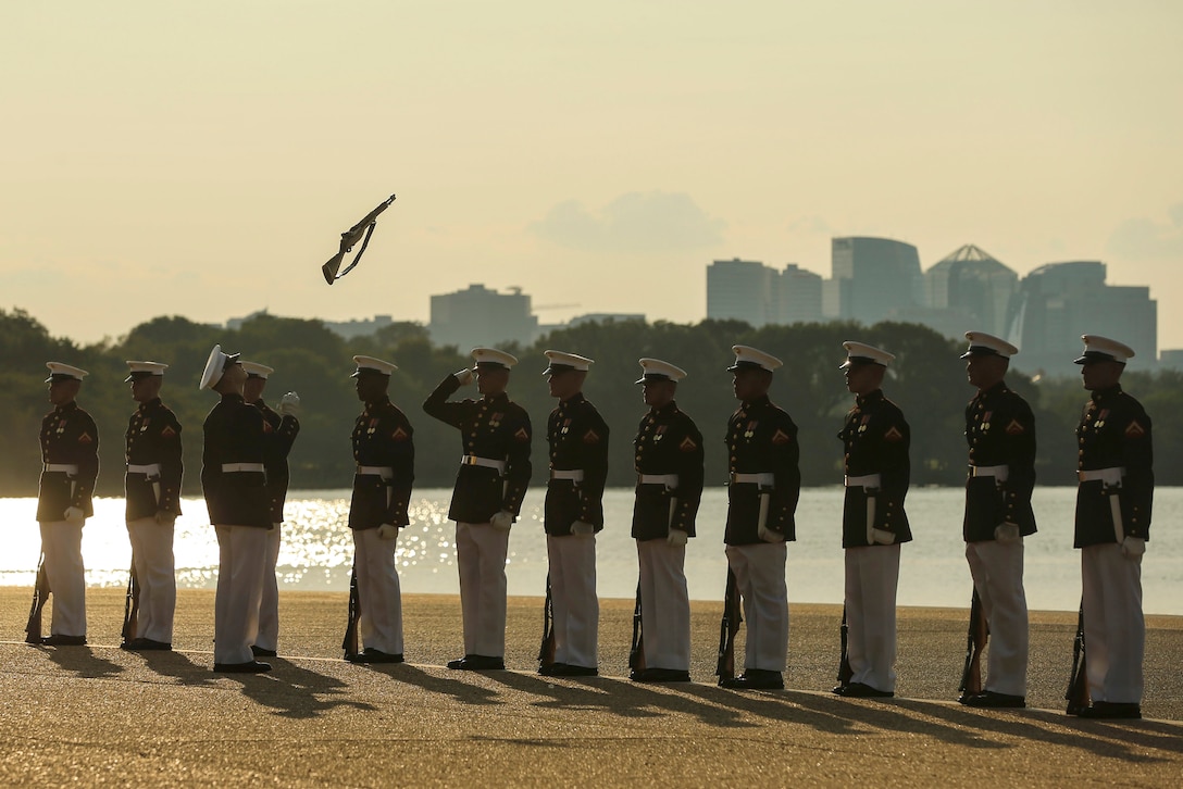 Marines stand in a line outside as one tosses a rifle in the air.