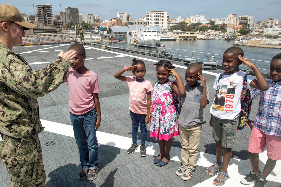 Kids show a sailor their practice salutes on a ship's deck.