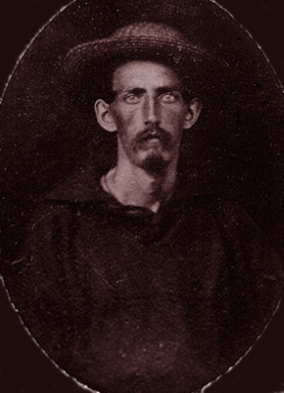 A man with a mustache wearing a wide-brimmed hat and Navy shirt looks at the camera.