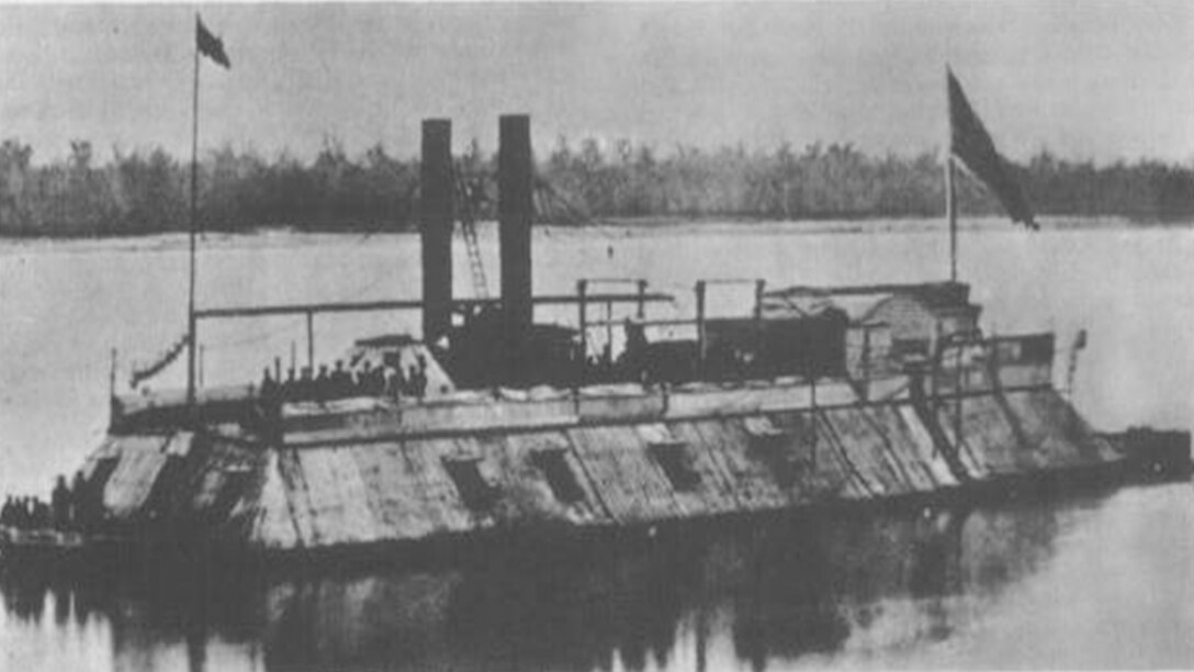 A ship covered in iron with two flags, two smokestacks and several sailors standing on it sails down a river.