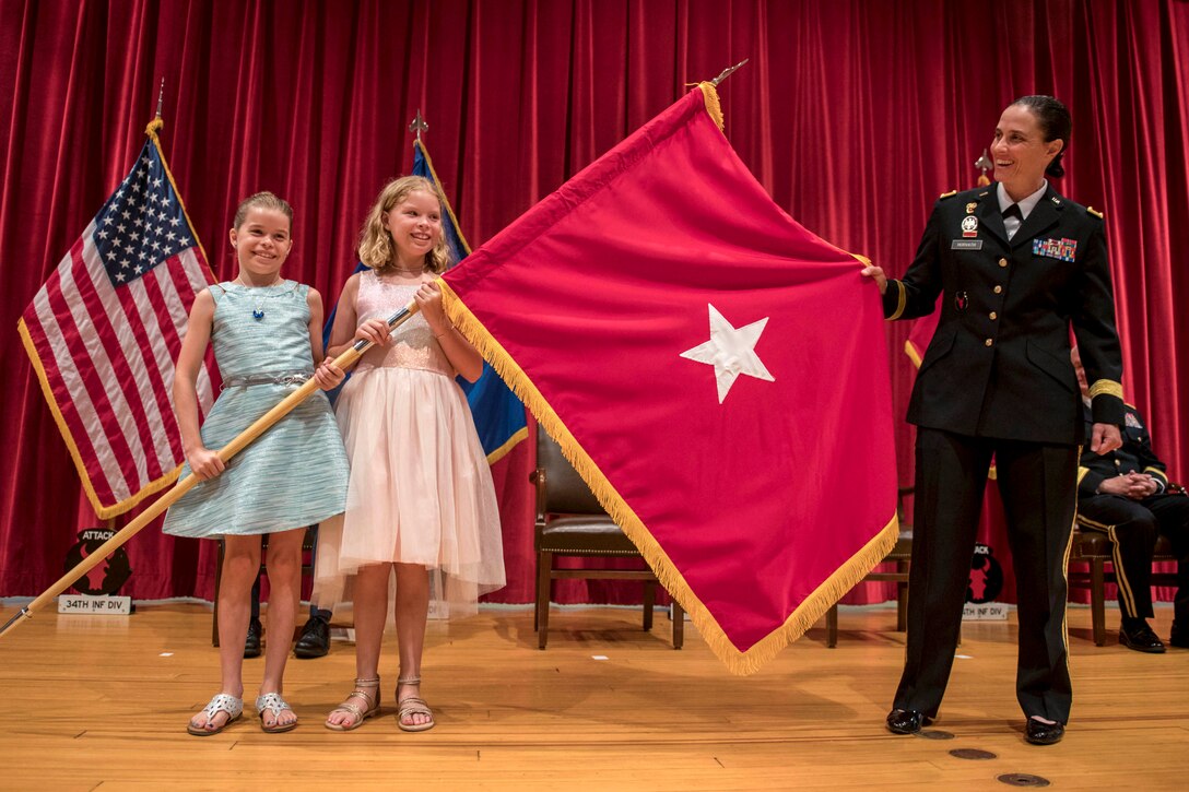 An Army general on a stage holds up a red flag with a white star in the center with help from her two smiling daughters.