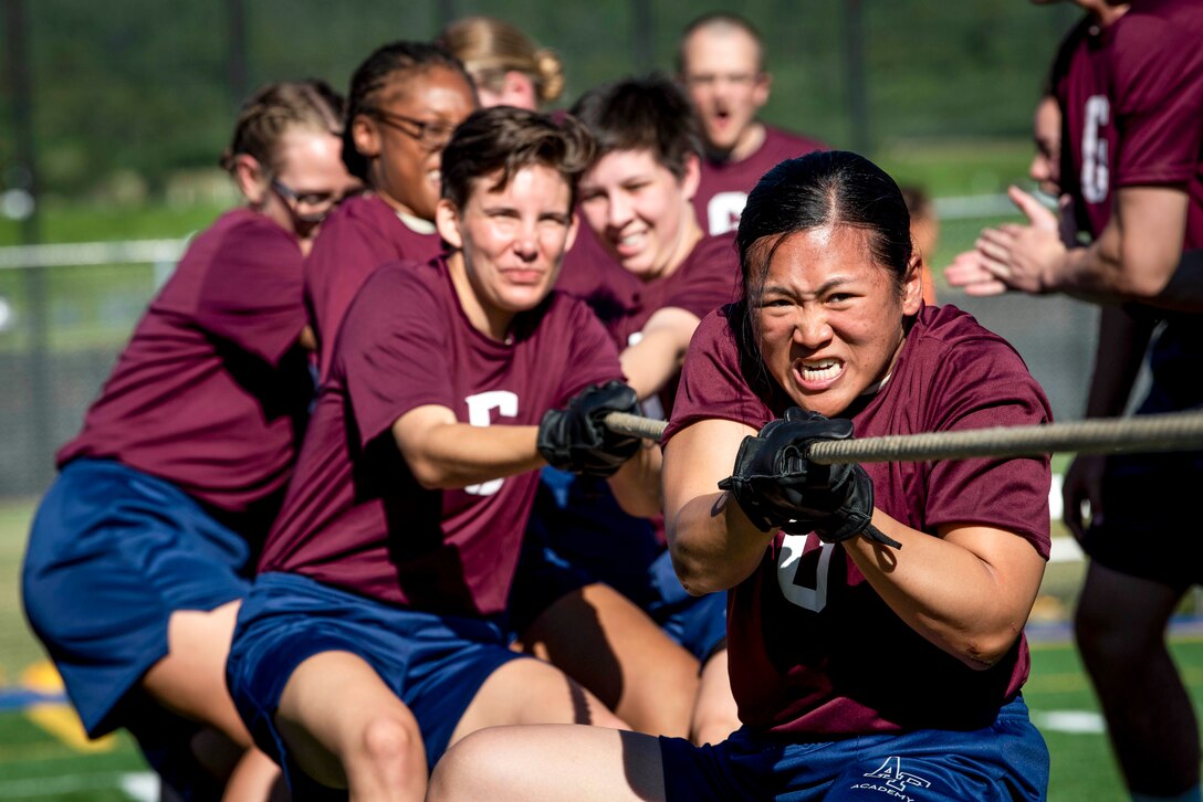 Air Force basic cadets in burgundy t-shirts and blue shorts pull a rope during tug of war outside.