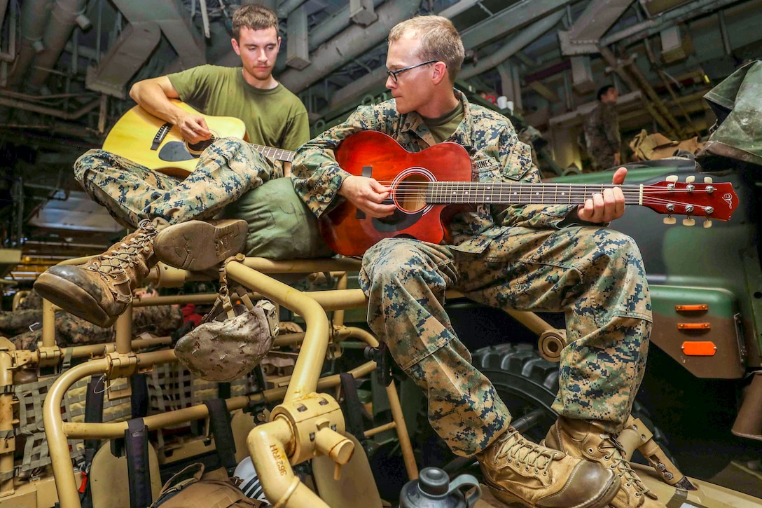 Two Marines sit on structural bars atop an open vehicle and play guitar.