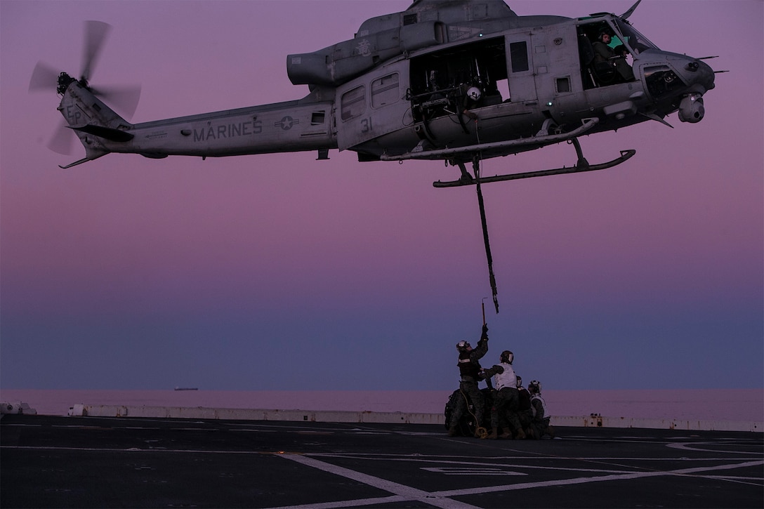 Marines stand below a helicopter during an exercise on a ship.