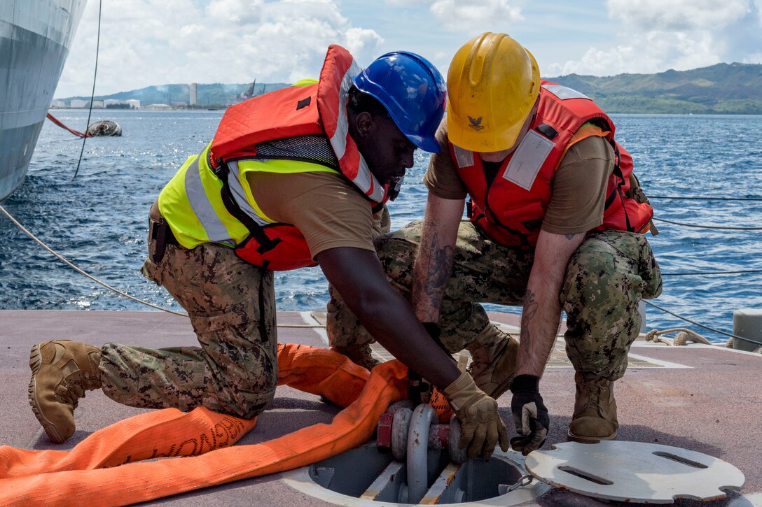 Two sailors work on lines on a pier near a ship.