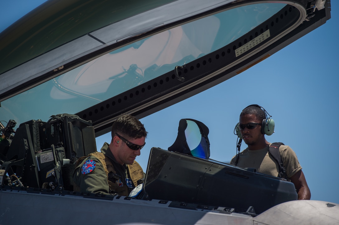An airman stands next to a pilot in the cockpit of an aircraft.