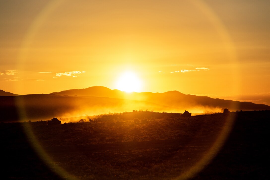 Military vehicles move along the ground as the sun rises behind some low hills.