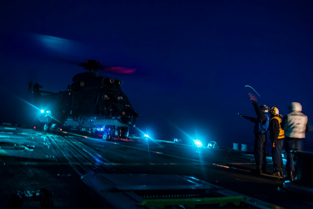 A helicopter parked at night with sailors standing off to the side.