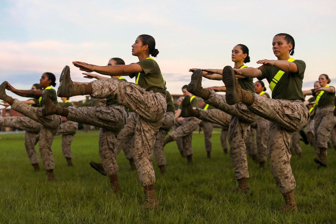 A group of Marines kick up one of their legs while reaching out their arms to touch their toes.