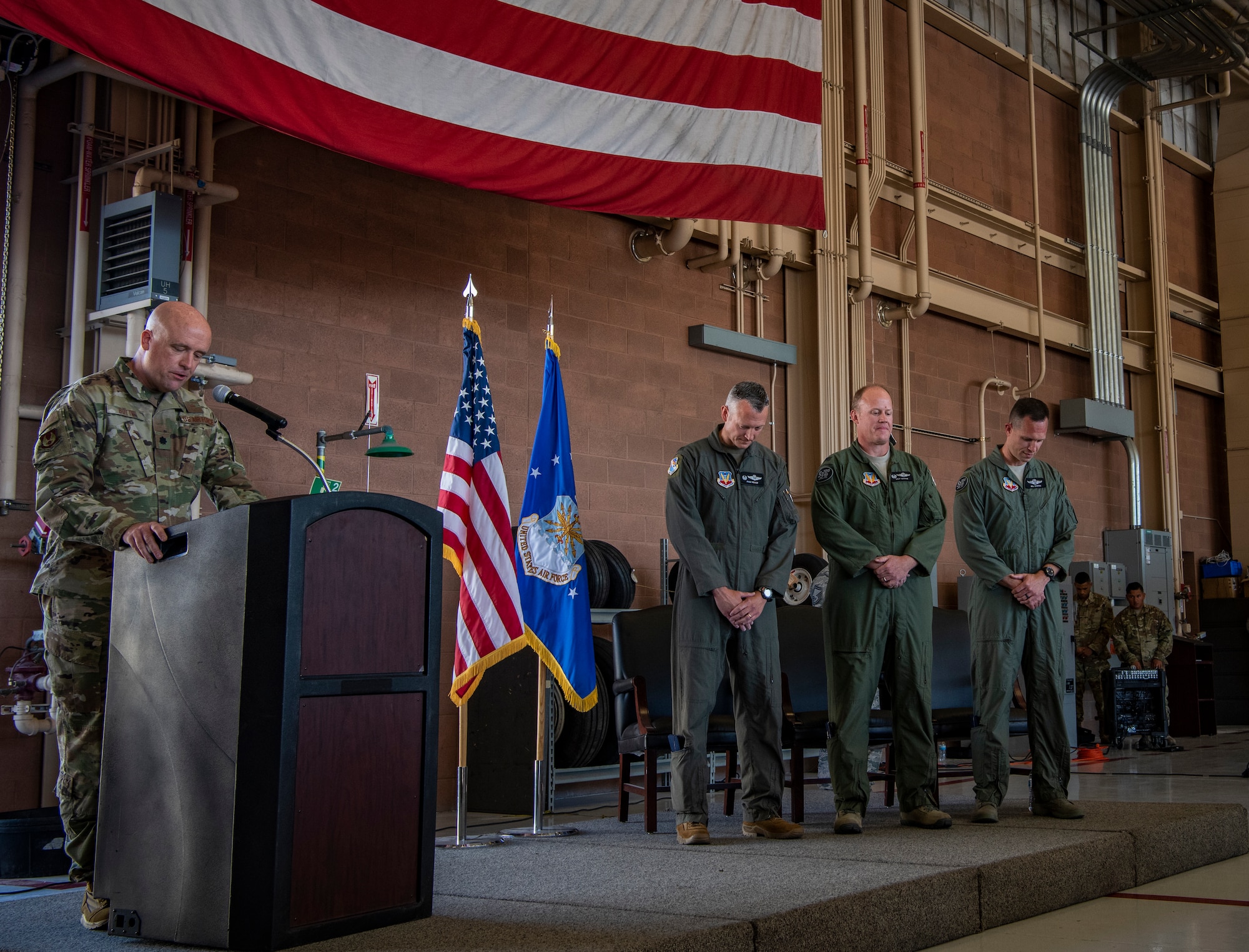 An Airman prays at a podium as three Airmen stand by on the stage.