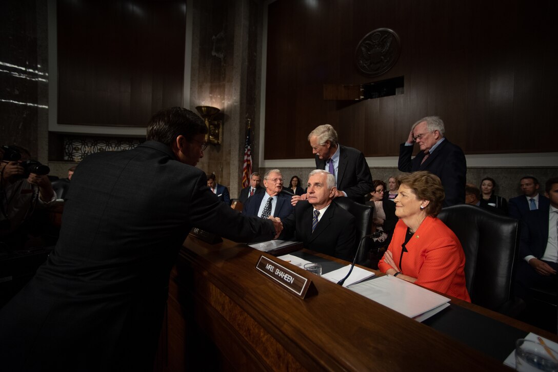 Several senators sit at a desk as a man reaches across to shake hands.