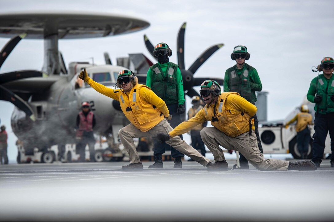 Two sailors kneel and point with other sailors and a  helicopter behind them.