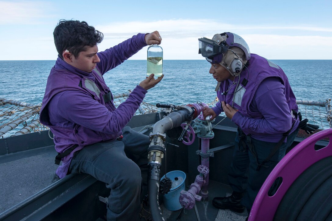 To  sailors hold up a fuel sample on a flight deck.