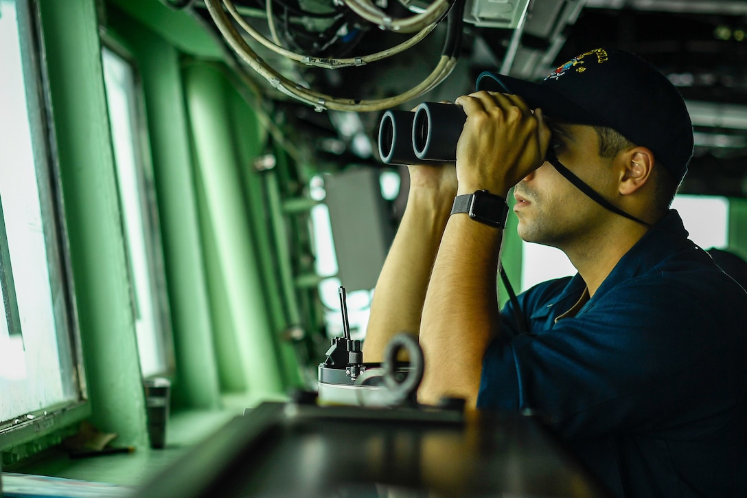 A seated sailor looks out a window using binoculars.
