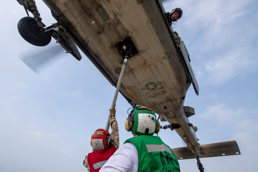 Two service members attach a hook to belly of hovering aircraft.