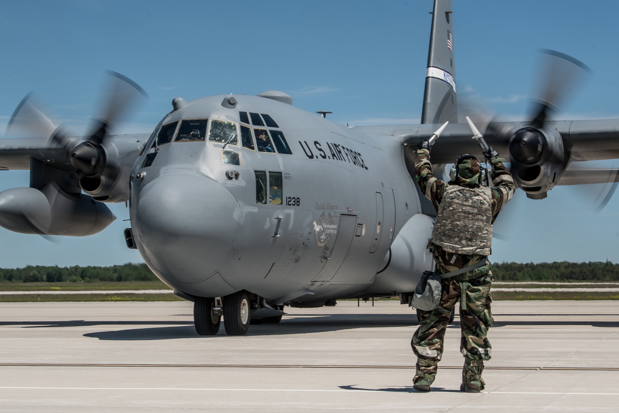 An aircraft maintainer wearing a chemical warfare defense ensemble directs a Kentucky Air National Guard C-130 Hercules aircraft to its parking spot at the Alpena Combat Readiness Training Center in Alpena, Mich., June 23, 2019, during exercise Charred Barrel. The event tested the wing’s ability to mobilize, fly to a remote site, and operate in a hostile environment. (U.S. Air National Guard photo by Staff Sgt. Joshua Horton)