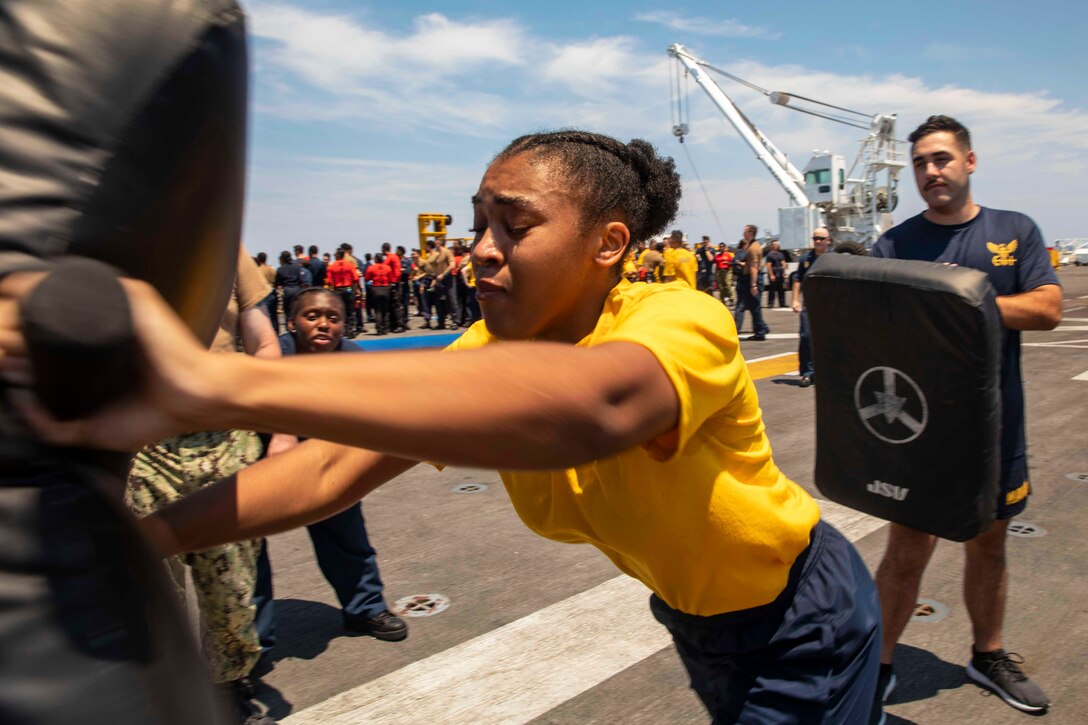 A sailor strikes a pad on deck of a ship while others look on.