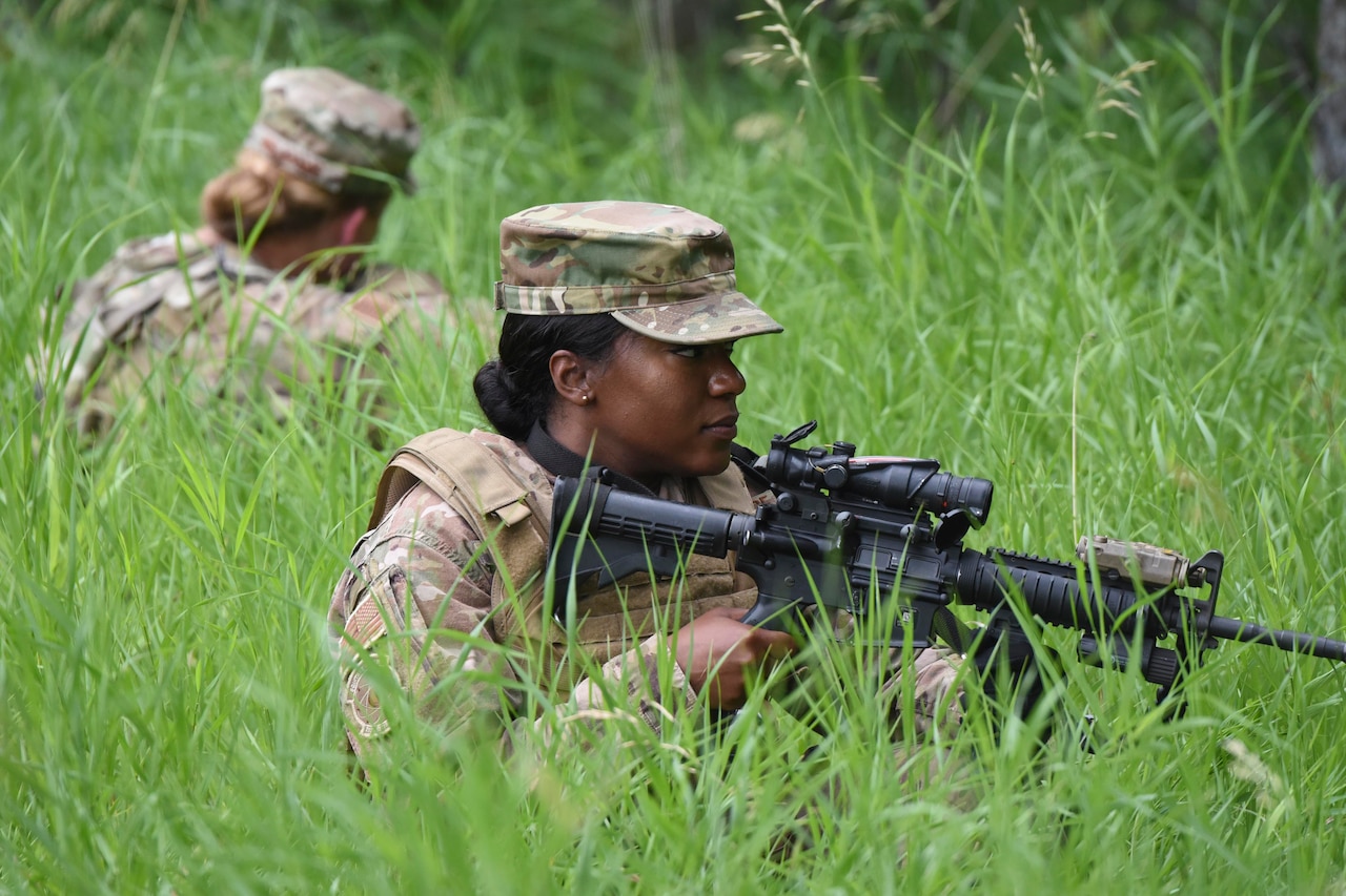 An airmen sits in the grass holding a weapon while another airmen sits behind
