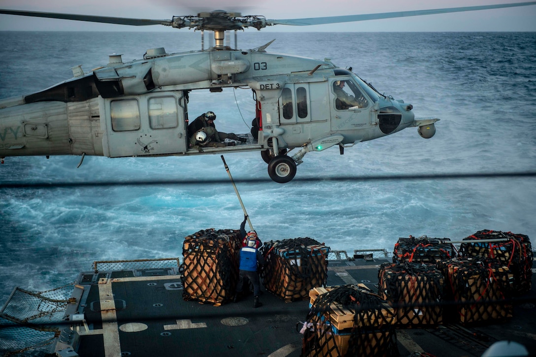 A sailor aboard holds out a pole to another sailor inside a helicopter.